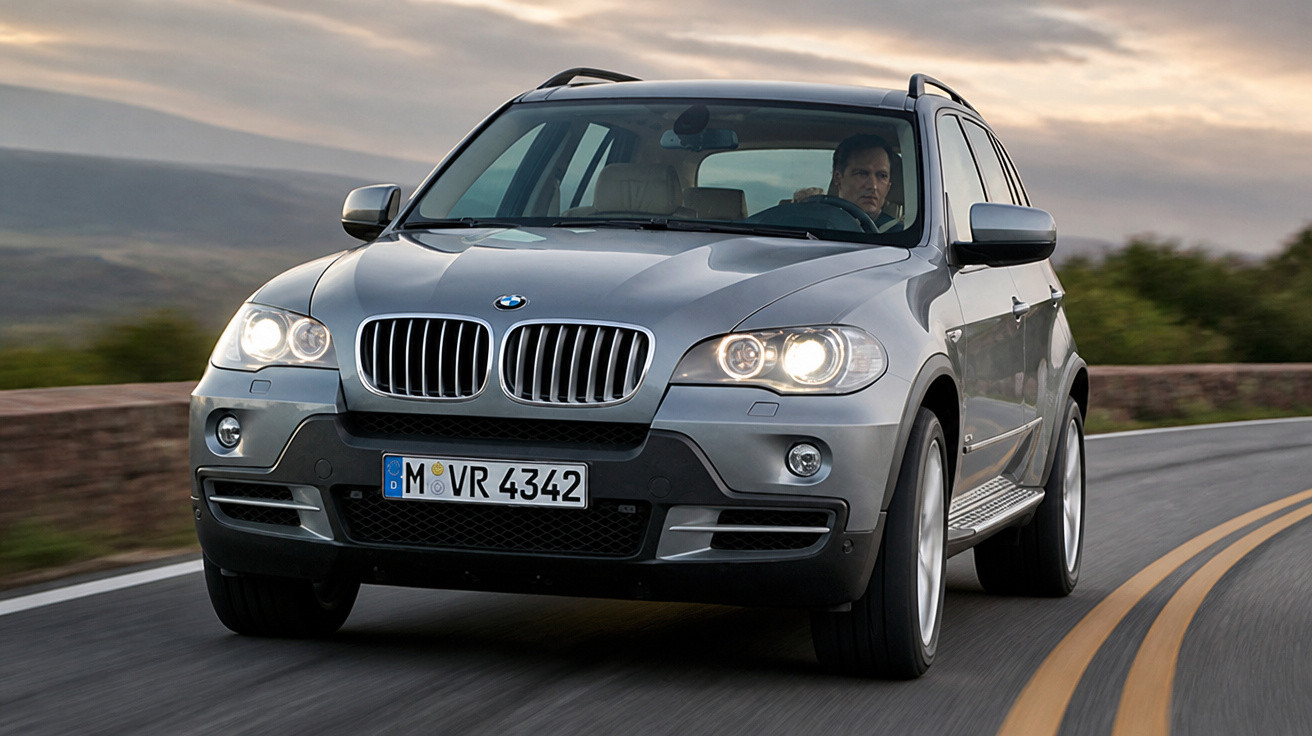 Silberner BMW SUV mit eingeschalteten Scheinwerfern fährt auf kurviger Landstrasse bei Dämmerung.