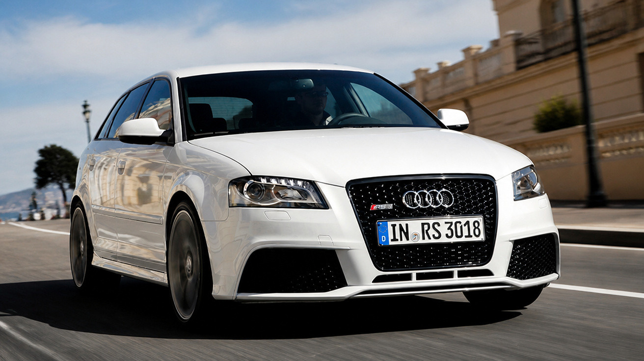 Weisser Audi RS3 fährt auf Strasse vor hellem Gebäude bei Tageslicht.